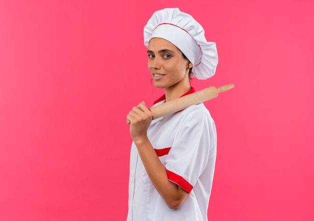Joven cocinera vistiendo uniforme de chef poniendo rodillo sobre el hombro en la pared rosa aislada con espacio de copia