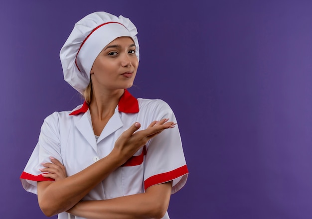 Joven cocinera vistiendo uniforme de chef puntos mano a lado en la pared aislada con espacio de copia