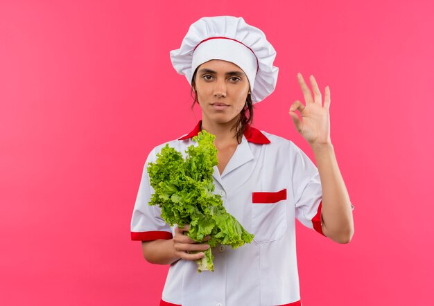 Joven cocinera vistiendo uniforme de chef sosteniendo ensalada y mostrando gesto okey en pared rosa aislada con espacio de copia