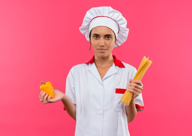 Joven cocinera vistiendo uniforme de chef sosteniendo espaguetis y papper en pared rosa aislada con espacio de copia