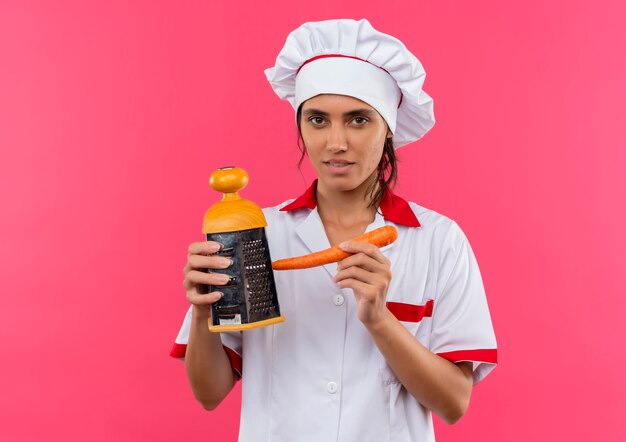 Joven cocinera vistiendo uniforme de chef sosteniendo rallado y zanahoria en pared rosa aislada con espacio de copia