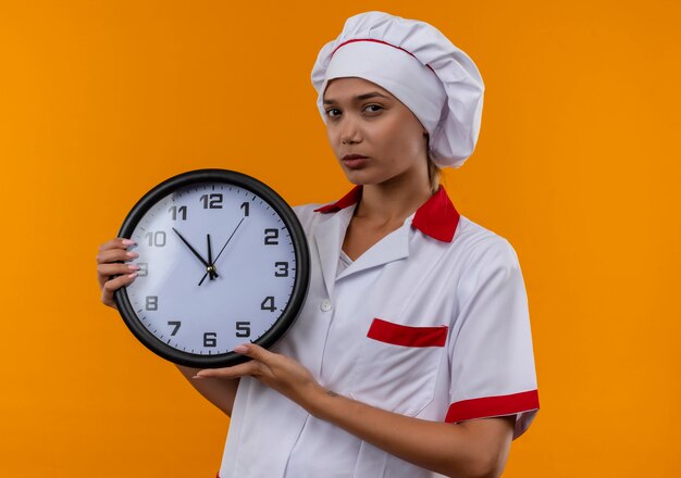 Joven cocinera vistiendo uniforme de chef sosteniendo el reloj de pared en la pared naranja aislada