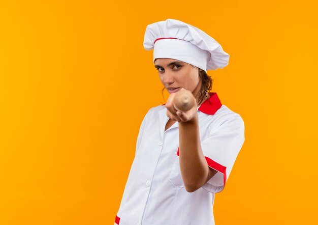 Joven cocinera vistiendo uniforme de chef sosteniendo el rodillo a la cámara en la pared amarilla aislada con espacio de copia