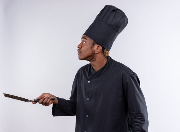 Joven cocinero afroamericano ansioso en uniforme de chef se coloca de lado y sostiene una sartén aislada sobre fondo blanco con espacio de copia