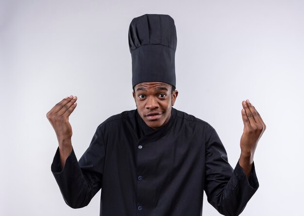 Joven cocinero afroamericano complacido en gestos de uniforme de chef espera signo de mano aislado en la pared blanca