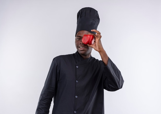 Joven cocinero afroamericano complacido en uniforme de chef cierra los ojos con pimiento rojo sobre blanco con espacio de copia