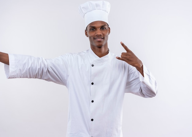 Joven cocinero afroamericano complacido en uniforme de chef hace gestos cuernos con la mano aislada en la pared blanca