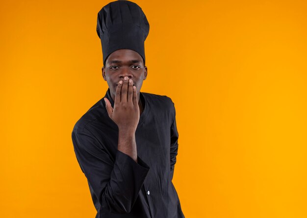 Joven cocinero afroamericano complacido en uniforme de chef pone la mano en la boca en naranja con espacio de copia