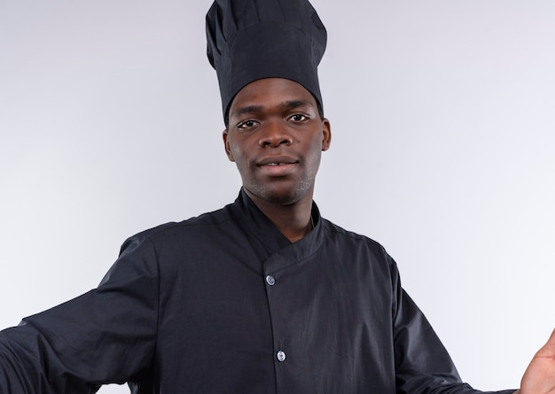 Joven cocinero afroamericano complacido en uniforme de chef pretende sostener la cámara aislada sobre fondo blanco con espacio de copia