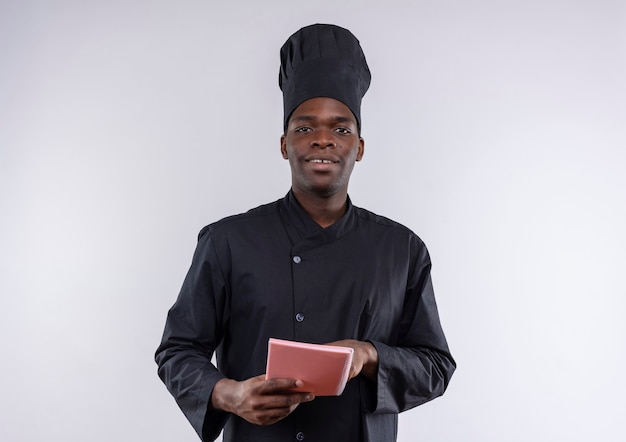 Joven cocinero afroamericano complacido en uniforme de chef sostiene y apunta en el cuaderno en blanco con espacio de copia