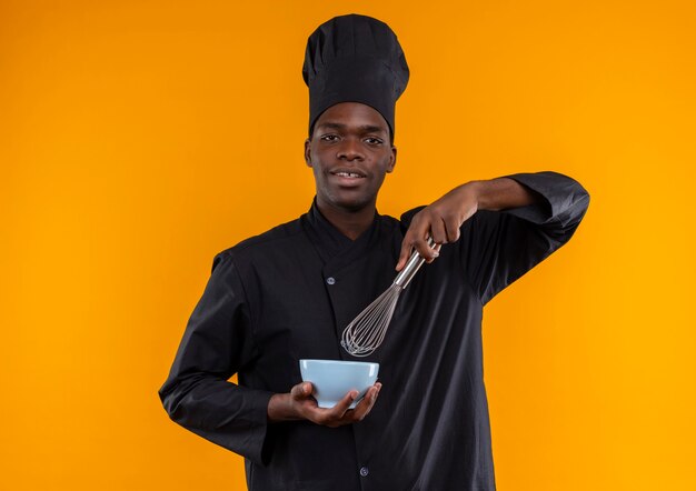 Joven cocinero afroamericano complacido en uniforme de chef sostiene un batidor y un tazón mirando a la cámara en naranja con espacio de copia