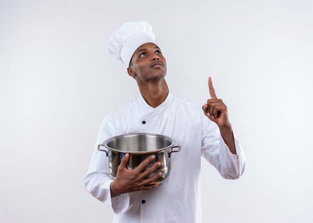 Joven cocinero afroamericano complacido en uniforme de chef sostiene una cacerola y apunta hacia arriba en la pared blanca aislada