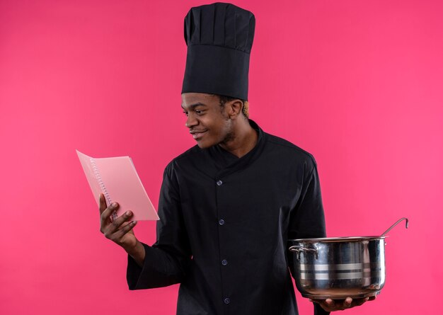 Joven cocinero afroamericano complacido en uniforme de chef sostiene una cacerola y un cuaderno aislado en la pared rosa