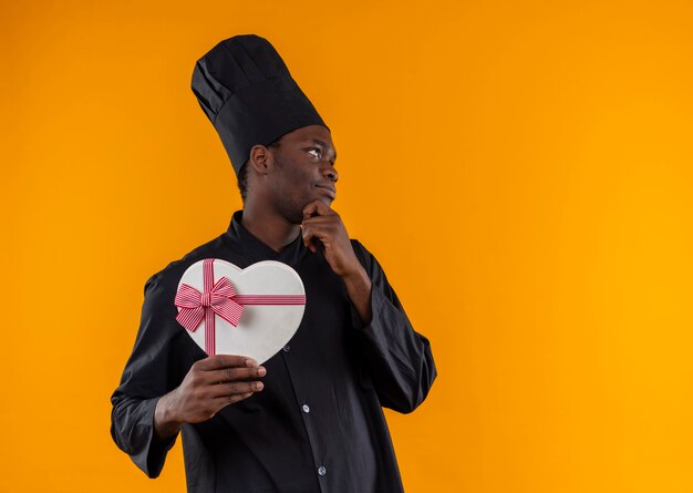 Joven cocinero afroamericano complacido en uniforme de chef sostiene una caja en forma de corazón y pone la mano en la barbilla en naranja con espacio de copia
