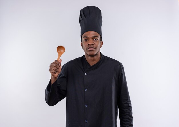 Joven cocinero afroamericano complacido en uniforme de chef sostiene una cuchara de madera aislada en la pared blanca