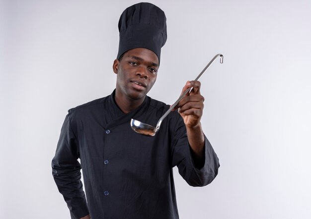 Joven cocinero afroamericano complacido en uniforme de chef sostiene cucharón en blanco con espacio de copia