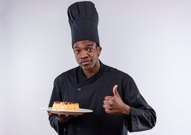 Joven cocinero afroamericano complacido en uniforme de chef sostiene pastel en plato y pulgar hacia arriba aislado en la pared blanca