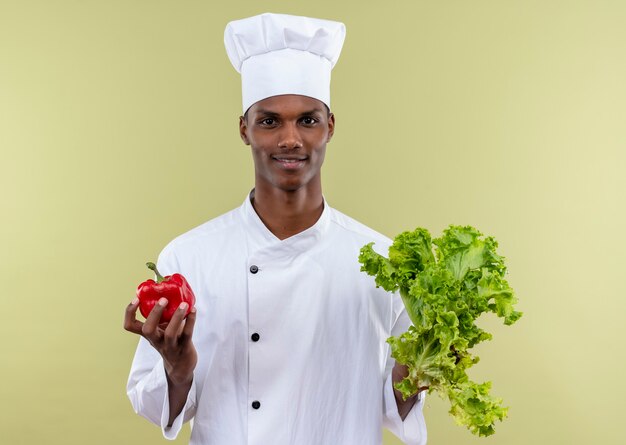 Joven cocinero afroamericano complacido en uniforme de chef sostiene pimiento rojo y ensalada aislado en la pared verde