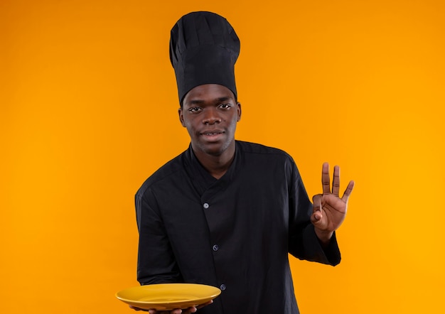 Joven cocinero afroamericano complacido en uniforme de chef sostiene placa y gestos ok signo de mano en naranja con espacio de copia