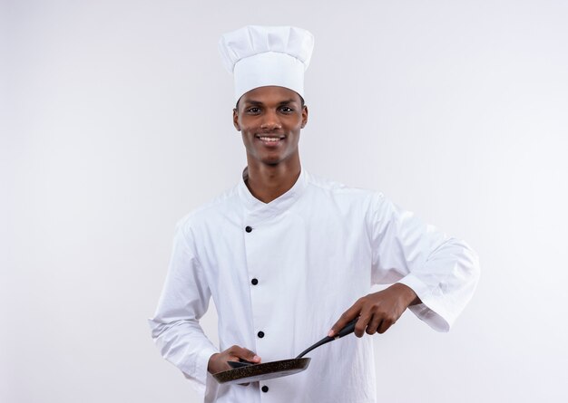 Joven cocinero afroamericano complacido en uniforme de chef sostiene una sartén y una cuchara en la pared blanca aislada