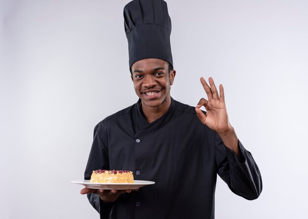 Joven cocinero afroamericano complacido en uniforme de chef sostiene la torta en el plato y gesticula el signo de la mano ok aislado en la pared blanca