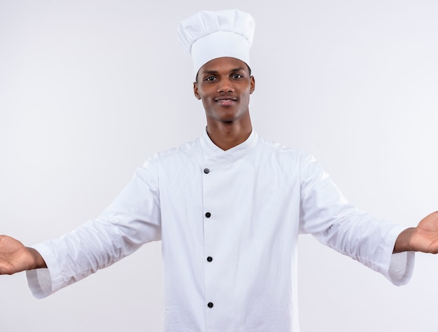 Joven cocinero afroamericano complacido en uniforme de chef tiene las manos abiertas aisladas en la pared blanca