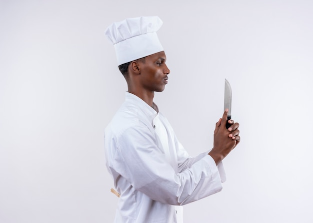 Joven cocinero afroamericano confiado en uniforme de chef se coloca de lado y sostiene un cuchillo como una pistola sobre fondo blanco aislado con espacio de copia
