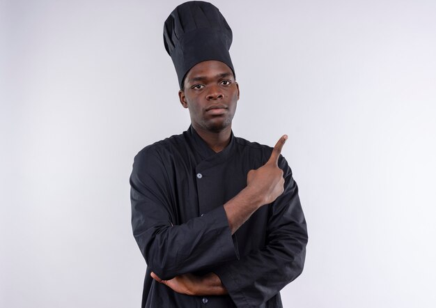 Joven cocinero afroamericano confiado en uniforme de chef cruza los brazos y apunta al lado en blanco con espacio de copia