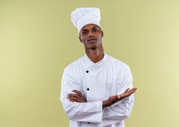 Joven cocinero afroamericano confiado en uniforme de chef cruza los brazos y mantiene la mano abierta aislada sobre fondo verde con espacio de copia