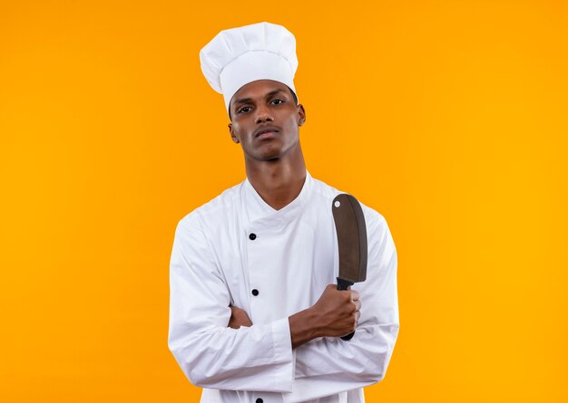 Joven cocinero afroamericano confiado en uniforme de chef cruza los brazos y sostiene un cuchillo aislado sobre fondo naranja con espacio de copia