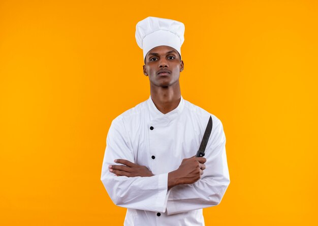 Joven cocinero afroamericano confiado en uniforme de chef cruza los brazos y sostiene un cuchillo aislado sobre fondo naranja con espacio de copia