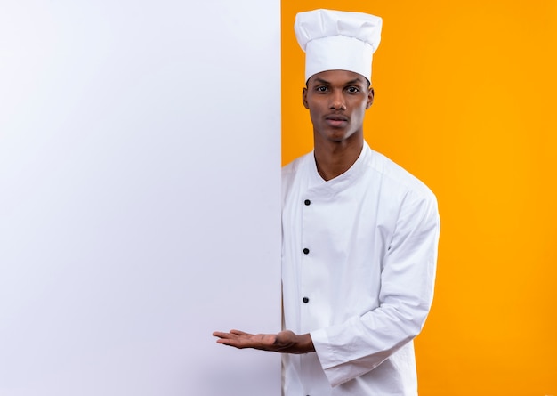 Joven cocinero afroamericano confiado en uniforme de chef está detrás de la pared blanca y apunta a la pared aislada sobre fondo naranja con espacio de copia