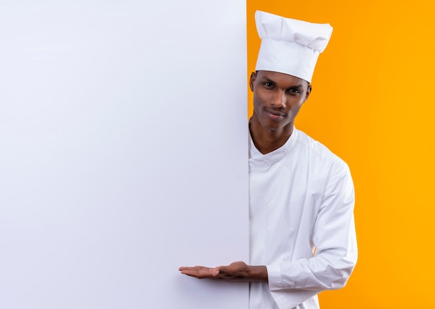 Joven cocinero afroamericano confiado en uniforme de chef está detrás de una pared blanca y sostiene la mano recta aislada sobre fondo naranja con espacio de copia