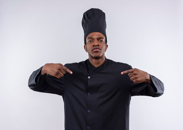 Joven cocinero afroamericano confiado en uniforme de chef se señala a sí mismo con ambas manos aisladas sobre fondo blanco con espacio de copia