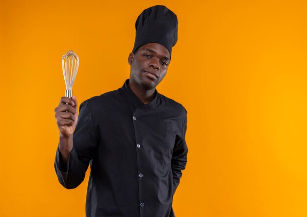 Joven cocinero afroamericano confiado en uniforme de chef sostiene batidor mirando a cámara aislada sobre fondo naranja con espacio de copia