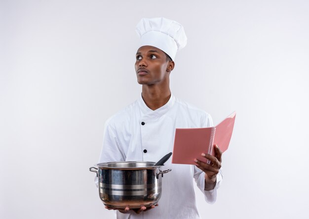 Joven cocinero afroamericano confiado en uniforme de chef sostiene una cacerola y un cuaderno mirando hacia el lado sobre fondo blanco aislado con espacio de copia