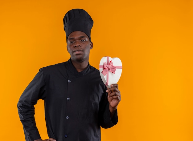 Joven cocinero afroamericano confiado en uniforme de chef sostiene una caja en forma de corazón y mira a la cámara en naranja con espacio de copia