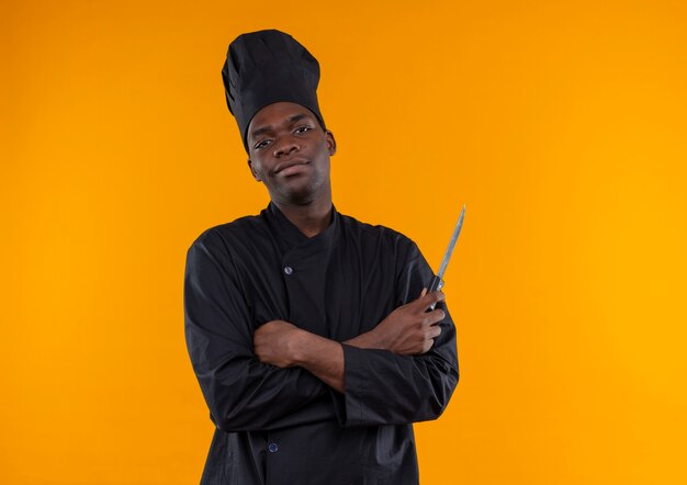 Joven cocinero afroamericano confiado en uniforme de chef sostiene un cuchillo con los brazos cruzados aislado sobre fondo naranja con espacio de copia