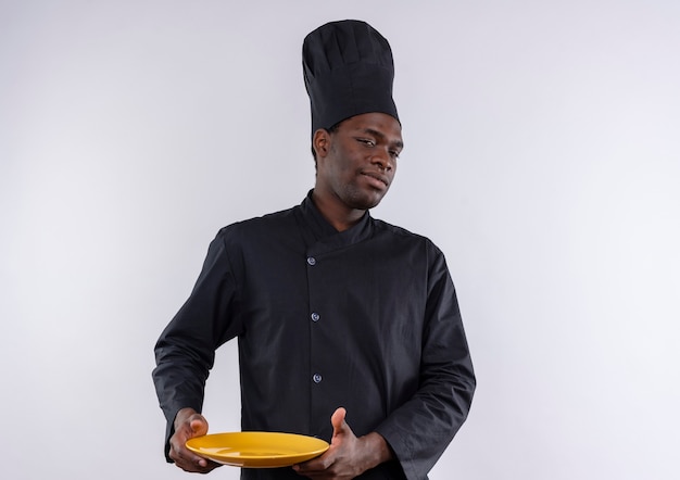 Joven cocinero afroamericano confiado en uniforme de chef sostiene la placa y mira a la cámara en blanco con espacio de copia