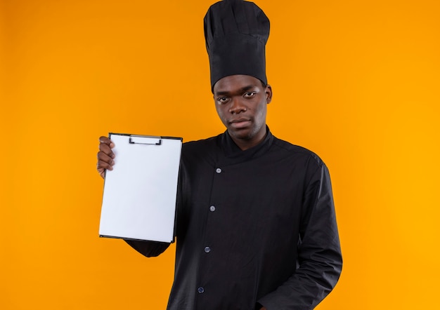 Joven cocinero afroamericano confiado en uniforme de chef sostiene el portapapeles en naranja con espacio de copia