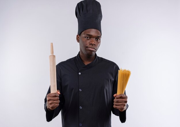 Joven cocinero afroamericano confiado en uniforme de chef sostiene un rodillo y un montón de espaguetis en blanco con espacio de copia
