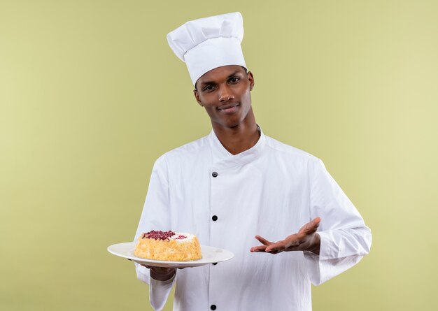 Joven cocinero afroamericano confiado en uniforme de chef sostiene la torta en el plato y puntos con la mano aislada sobre fondo verde con espacio de copia