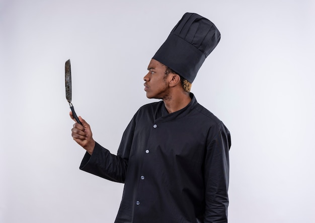 Foto gratuita joven cocinero afroamericano confundido con uniforme de chef se coloca de lado y sostiene sartén aislado en la pared blanca
