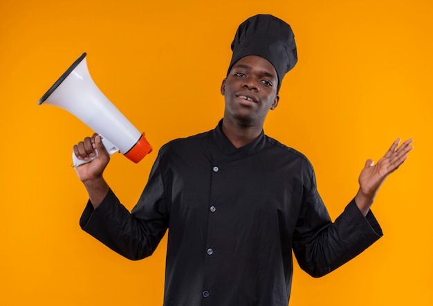 Joven cocinero afroamericano confundido en uniforme de chef sostiene altavoz y levanta la mano en naranja con espacio de copia