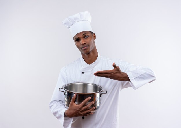 Joven cocinero afroamericano confundido en uniforme de chef sostiene y apunta a una cacerola en la pared blanca aislada