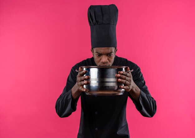 Joven cocinero afroamericano confundido en uniforme de chef sostiene y mira una cacerola aislada en la pared rosa