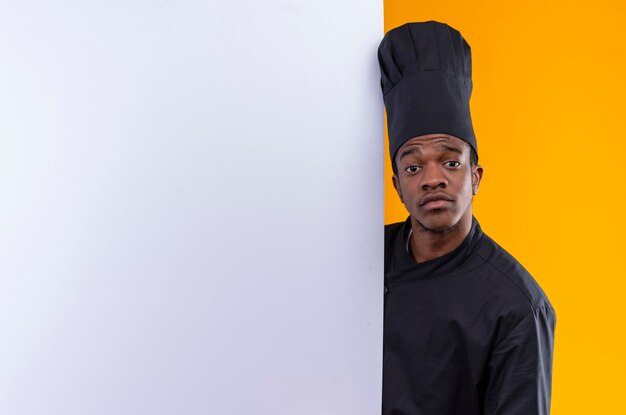 Joven cocinero afroamericano molesto en uniforme de chef está detrás de una pared blanca aislada sobre fondo naranja con espacio de copia