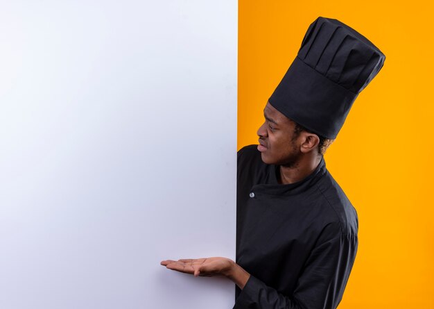 Joven cocinero afroamericano molesto en uniforme de chef está detrás de la pared blanca y apunta a la pared aislada sobre fondo naranja con espacio de copia