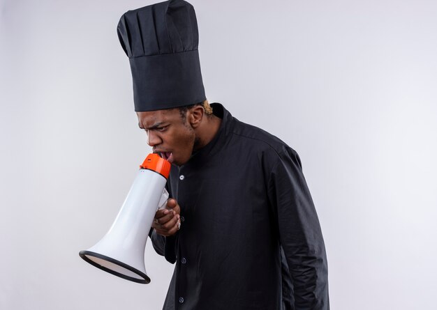 Joven cocinero afroamericano molesto en uniforme de chef grita a través del altavoz aislado sobre fondo blanco con espacio de copia