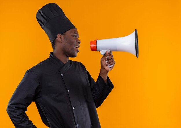 Joven cocinero afroamericano molesto con uniforme de chef se para de lado y habla a través del altavoz en naranja con espacio de copia